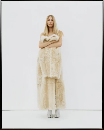 Fashion shot of a blonde woman posing indoors in a large beige fur coat, wearing beige boots with a white backdrop.