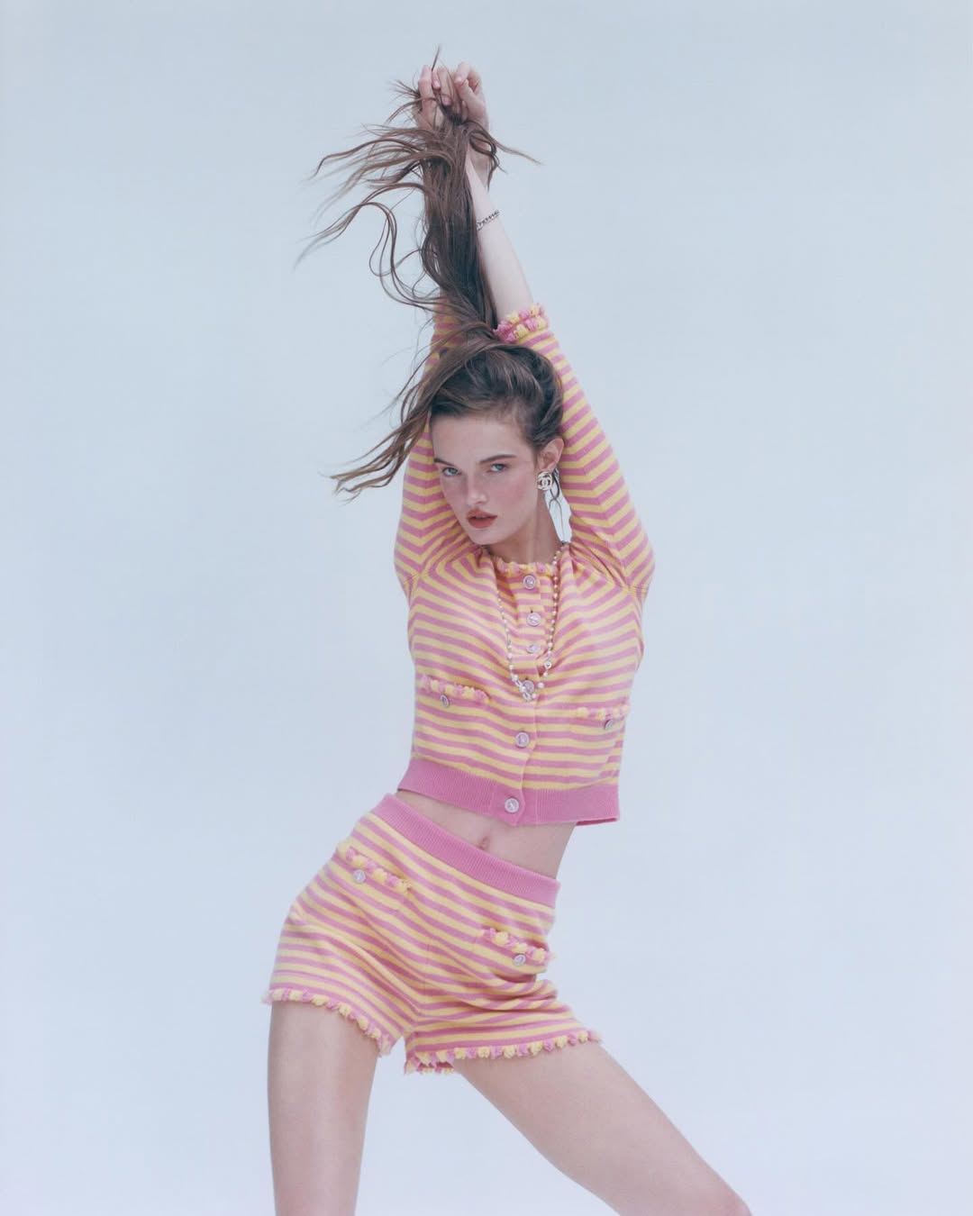 A young woman in a pink and peach striped two-piece set poses with her arms raised, hair flowing, against a light backdrop in a studio shot.
