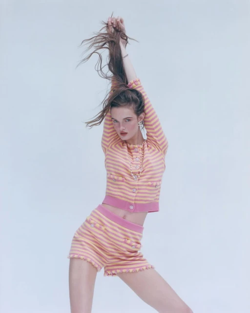 A young woman in a pink and peach striped two-piece set poses with her arms raised, hair flowing, against a light backdrop in a studio shot.