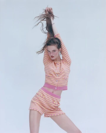 A young woman in a pink and peach striped two-piece set poses with her arms raised, hair flowing, against a light backdrop in a studio shot.