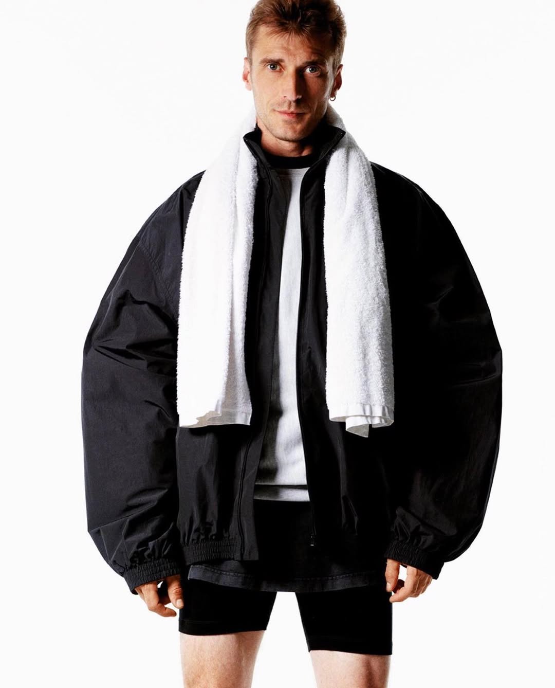 Athletic man with a towel around his neck wearing a black jacket and shorts posing against a bright white background in a studio setting.