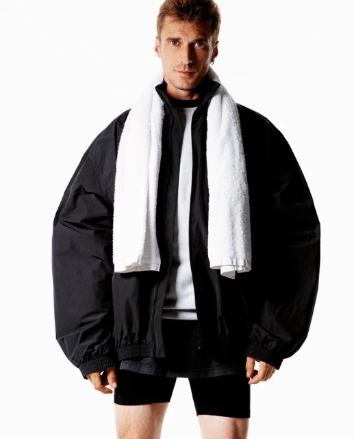 Athletic man with a towel around his neck wearing a black jacket and shorts posing against a bright white background in a studio setting.