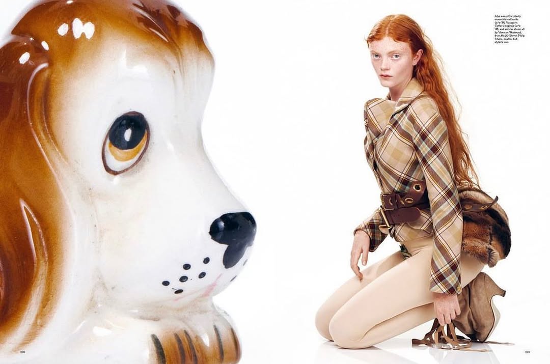 Woman in plaid blazer poses on knees near ceramic dog head in a fashion shoot on a white backdrop in a studio setting.