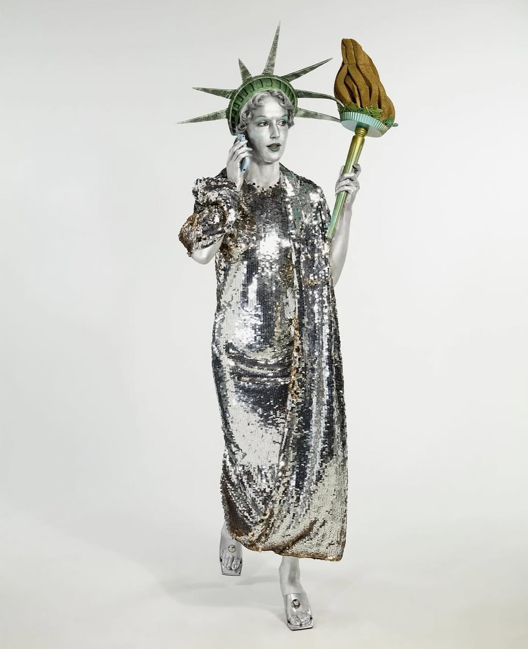 Woman in Statue of Liberty costume holds a phone, wearing a shimmering silver dress and a torch. Studio shot, light background.