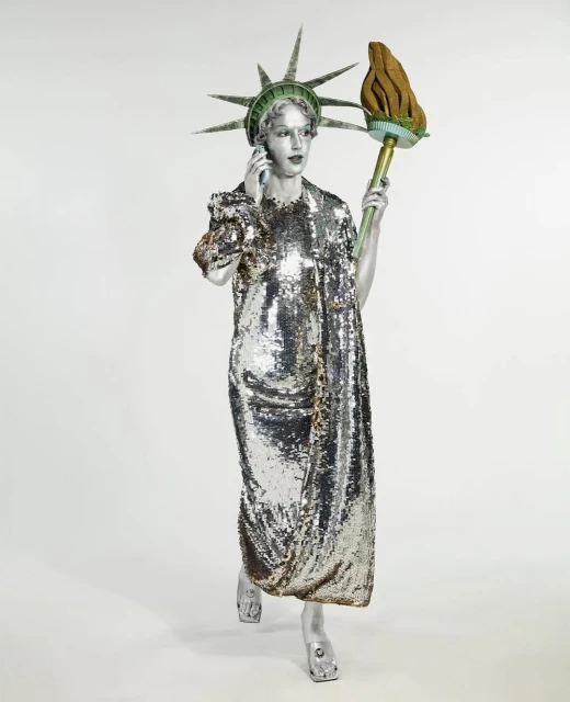 Woman in Statue of Liberty costume holds a phone, wearing a shimmering silver dress and a torch. Studio shot, light background.