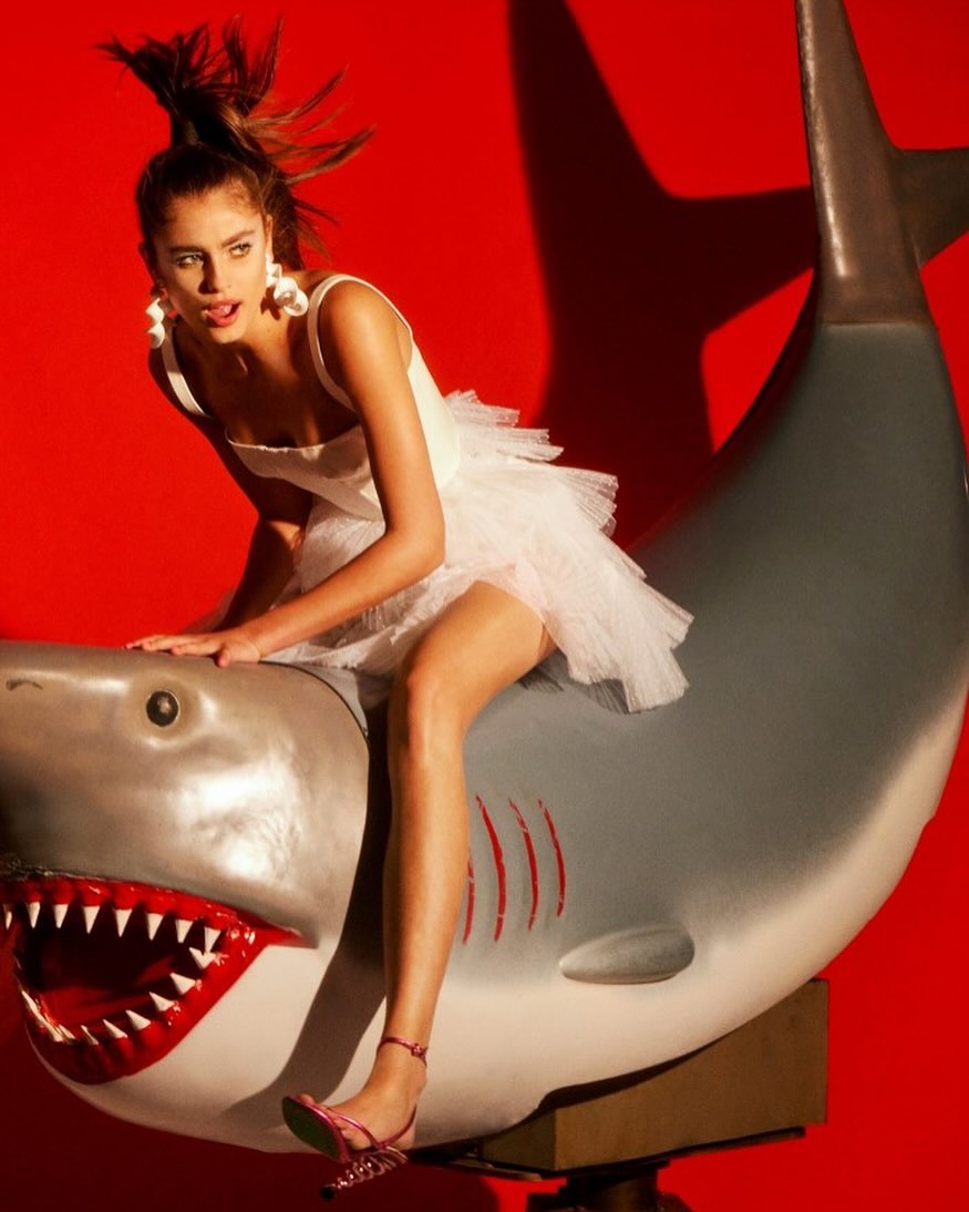 A woman in a white dress rides a gray shark prop against a red background, creating a striking and whimsical fashion statement.