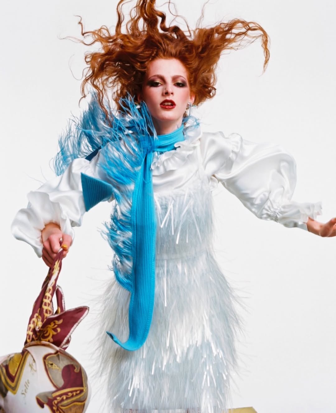 A red-haired fashion model in a white frilly dress, accessorized with a blue scarf, poses against a clean white backdrop with a patterned bag.