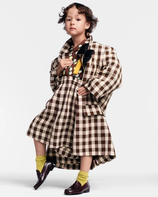Young child poses in a brown and white plaid oversized suit with dark brown loafers and yellow socks against a white backdrop.