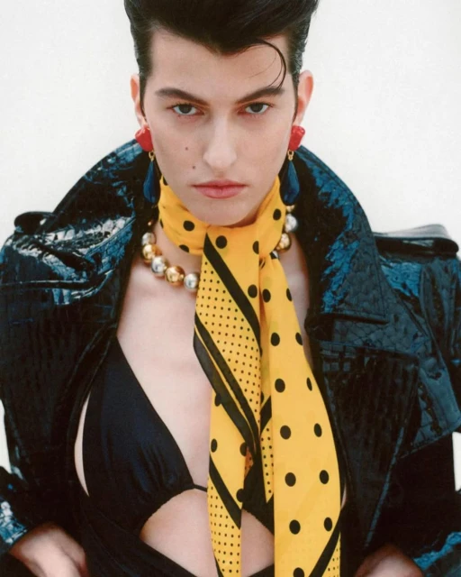 Fashionable person poses in a black bikini, black jacket, yellow scarf, and jewelry against a white backdrop in a studio setting.