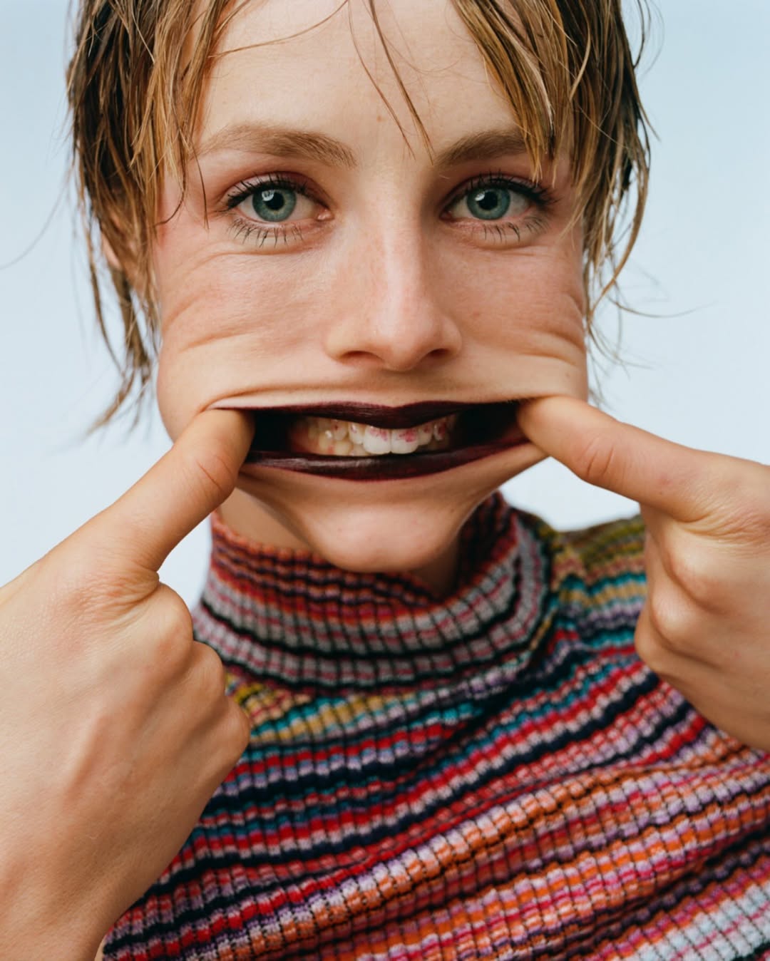 Close-up portrait of a woman stretching her face skin with her fingers, showing teeth, wearing a colourful striped top indoors.