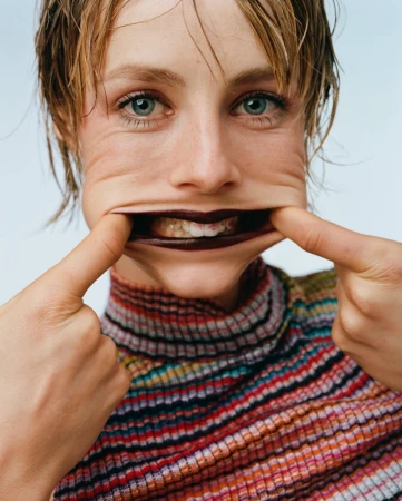 Close-up portrait of a woman stretching her face skin with her fingers, showing teeth, wearing a colourful striped top indoors.