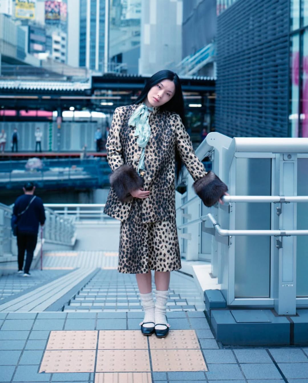 Asian woman in leopard print suit poses on stairs in an urban setting with tall buildings. She has long black hair and fur cuffs.