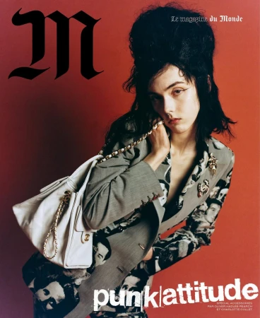 A woman with dark teased hair wearing a patterned suit and holding a white chain-strap handbag against a red background.
