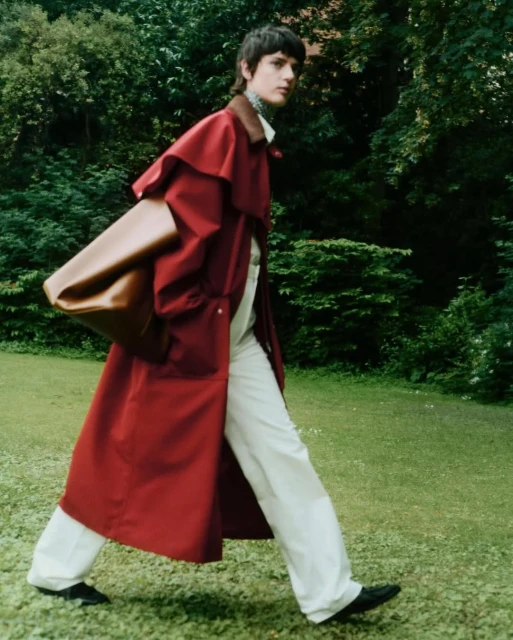 A brunette adult wearing a red trench coat and white pants walks across a green lawn with leafy trees in the background, carrying a large brown bag.