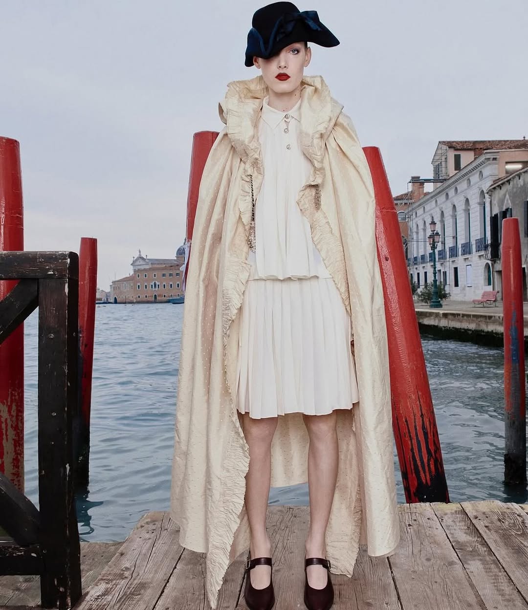 Woman poses in cape and hat on weathered dock in Venice, Italy, with waterfront buildings and canal visible in background. Fashion shoot.