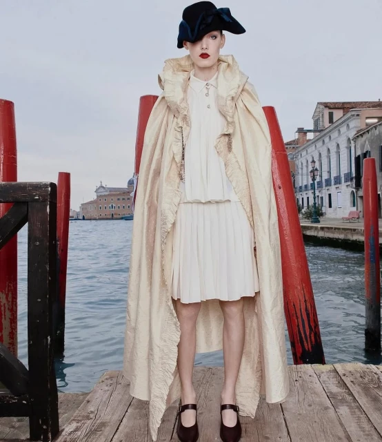 Woman poses in cape and hat on weathered dock in Venice, Italy, with waterfront buildings and canal visible in background. Fashion shoot.