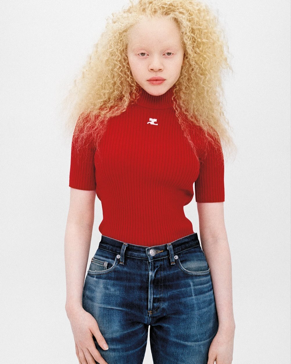 Albino fashion model poses in red short-sleeve top and jeans against a white backdrop. The model has pale skin and blonde curly hair.