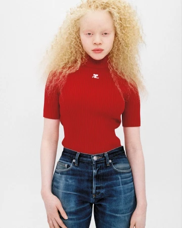 Albino fashion model poses in red short-sleeve top and jeans against a white backdrop. The model has pale skin and blonde curly hair.