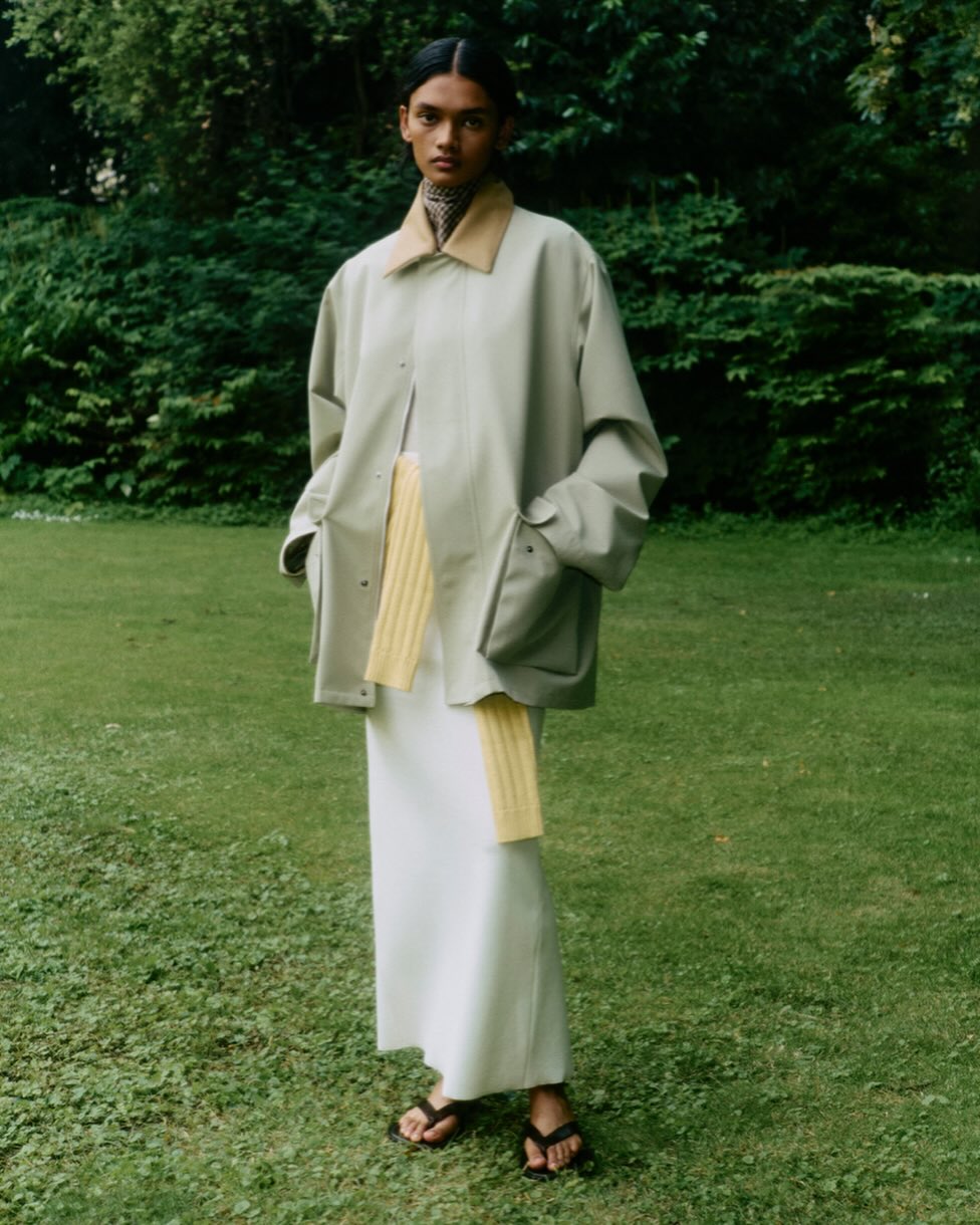 Model standing on grass wearing an oversized light green jacket, yellow scarf, white skirt, and black sandals