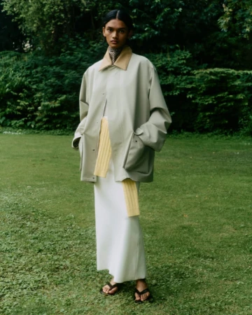 Model standing on grass wearing an oversized light green jacket, yellow scarf, white skirt, and black sandals