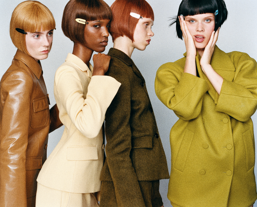Four women with short haircuts and hair clips pose in studio wearing color-coordinated, tailored suit jackets and skirts.