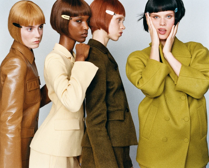 Four women with short haircuts and hair clips pose in studio wearing color-coordinated, tailored suit jackets and skirts.