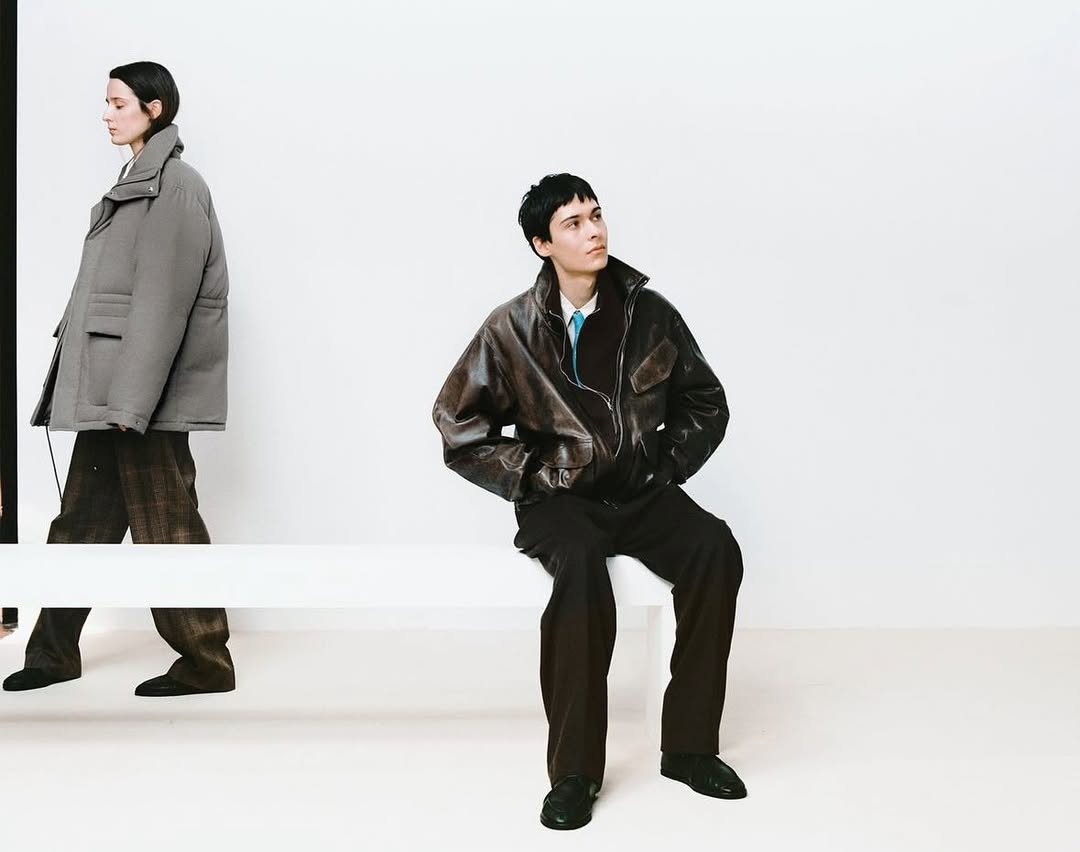 Two fashion models pose in an indoor setting against a bright white wall. One model is walking while the other is seated on a bench.