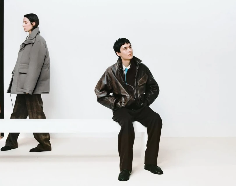 Two fashion models pose in an indoor setting against a bright white wall. One model is walking while the other is seated on a bench.