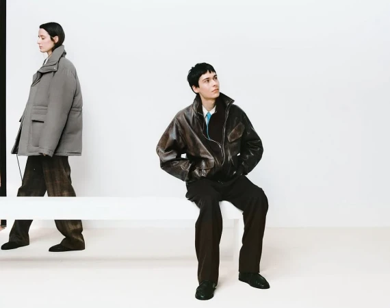 Two fashion models pose in an indoor setting against a bright white wall. One model is walking while the other is seated on a bench.
