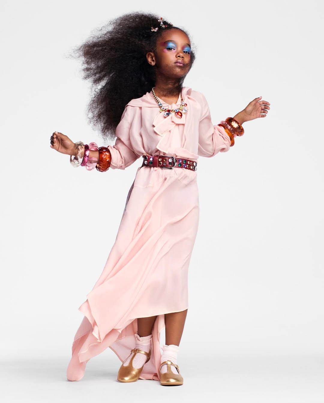Portrait of a child with dark hair in a flowing pink dress, statement jewelry, and gold shoes, posing on a white background.