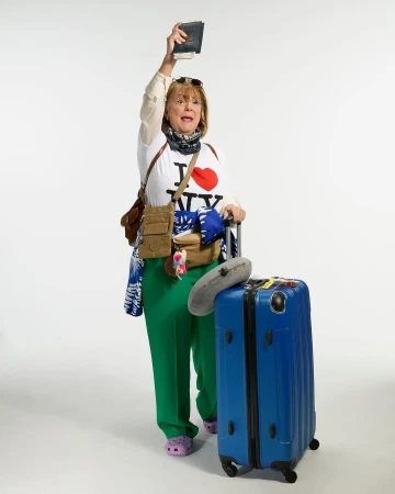 Full shot of a mature woman in travel attire, holding a passport, standing with a suitcase, expressing excitement on a white background.