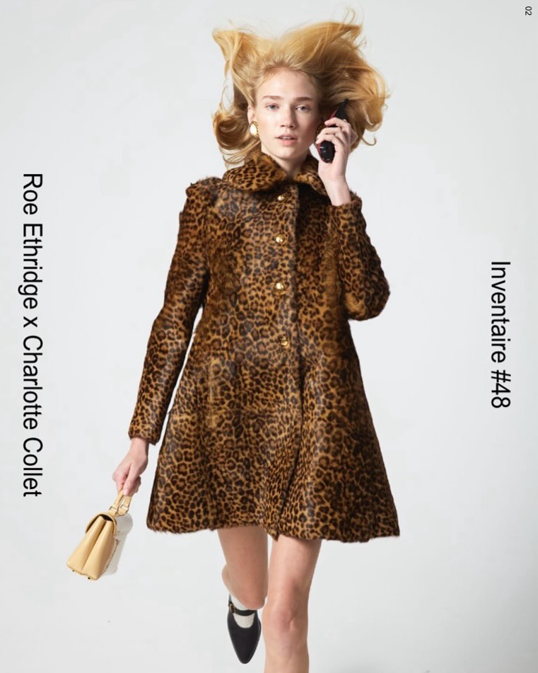 A woman in a leopard print coat holds a phone and purse against a white backdrop, showcasing fashion and style in a studio setting.