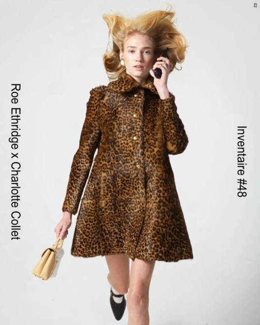 A woman in a leopard print coat holds a phone and purse against a white backdrop, showcasing fashion and style in a studio setting.