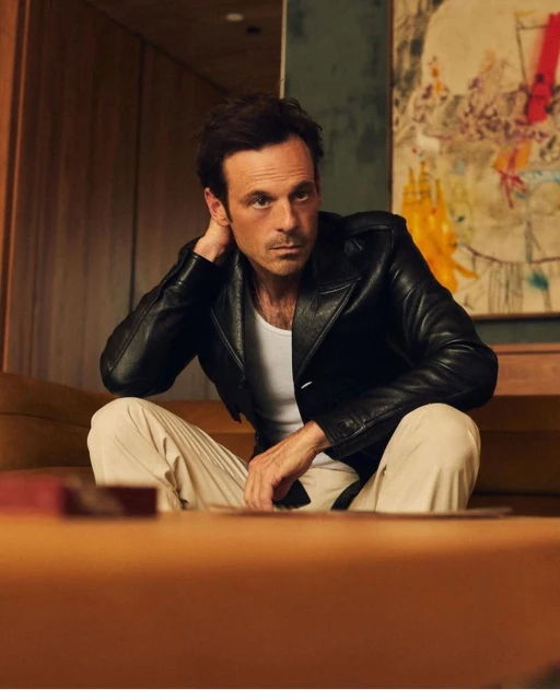 A brunette adult male in a black jacket and cream pants sits indoors, against a neutral backdrop with a painting in the background.