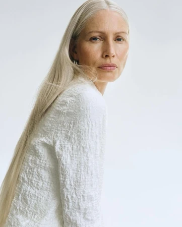 Mature woman with long straight grey hair wearing a textured white top against a clean white background
