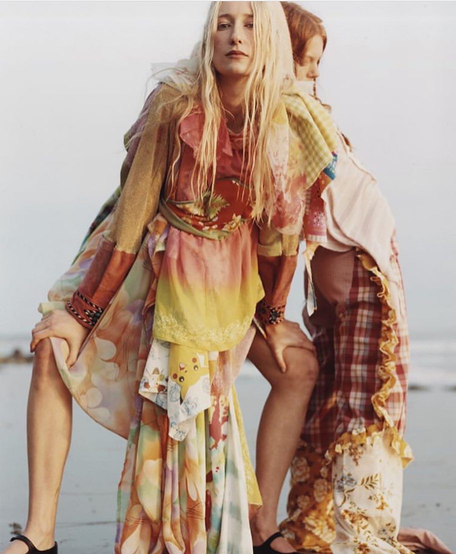 Two fashion models in flowing dresses pose back-to-back on a sandy beach with a light sky. Bohemian aesthetic.