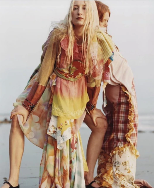 Two fashion models in flowing dresses pose back-to-back on a sandy beach with a light sky. Bohemian aesthetic.