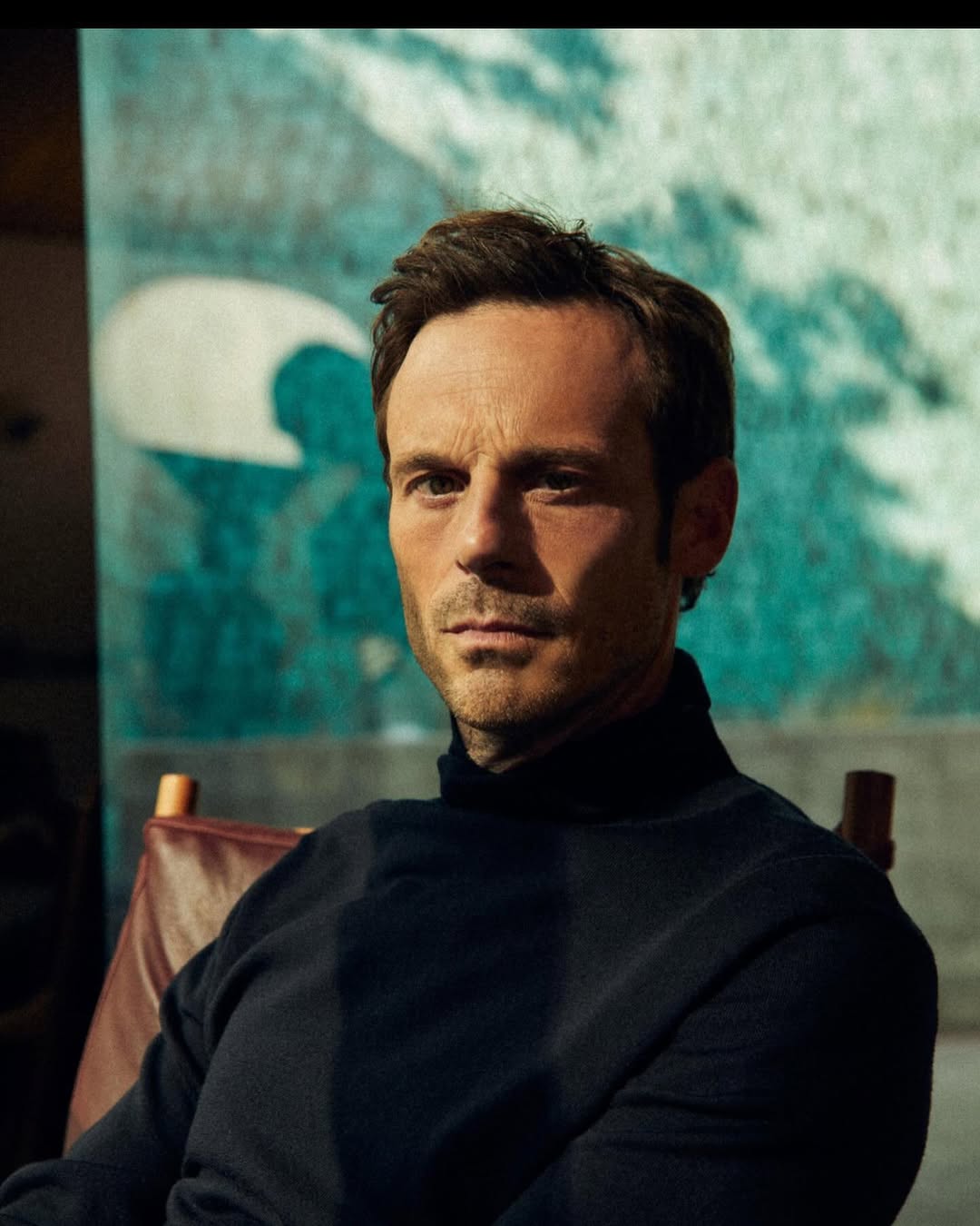 Portrait of an adult male sitting indoors. He is wearing a black turtleneck sweater and is posed in front of a turquoise backdrop.
