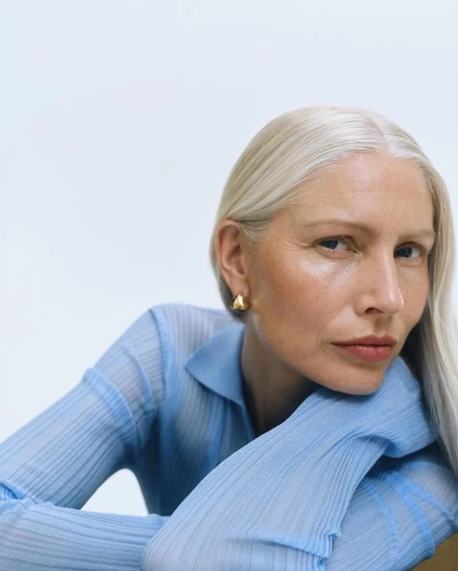 Close-up portrait of a mature woman with white hair, wearing a light blue top and gold earrings, resting her head on her hands.