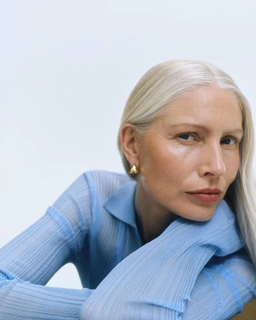 Close-up portrait of a mature woman with white hair, wearing a light blue top and gold earrings, resting her head on her hands.