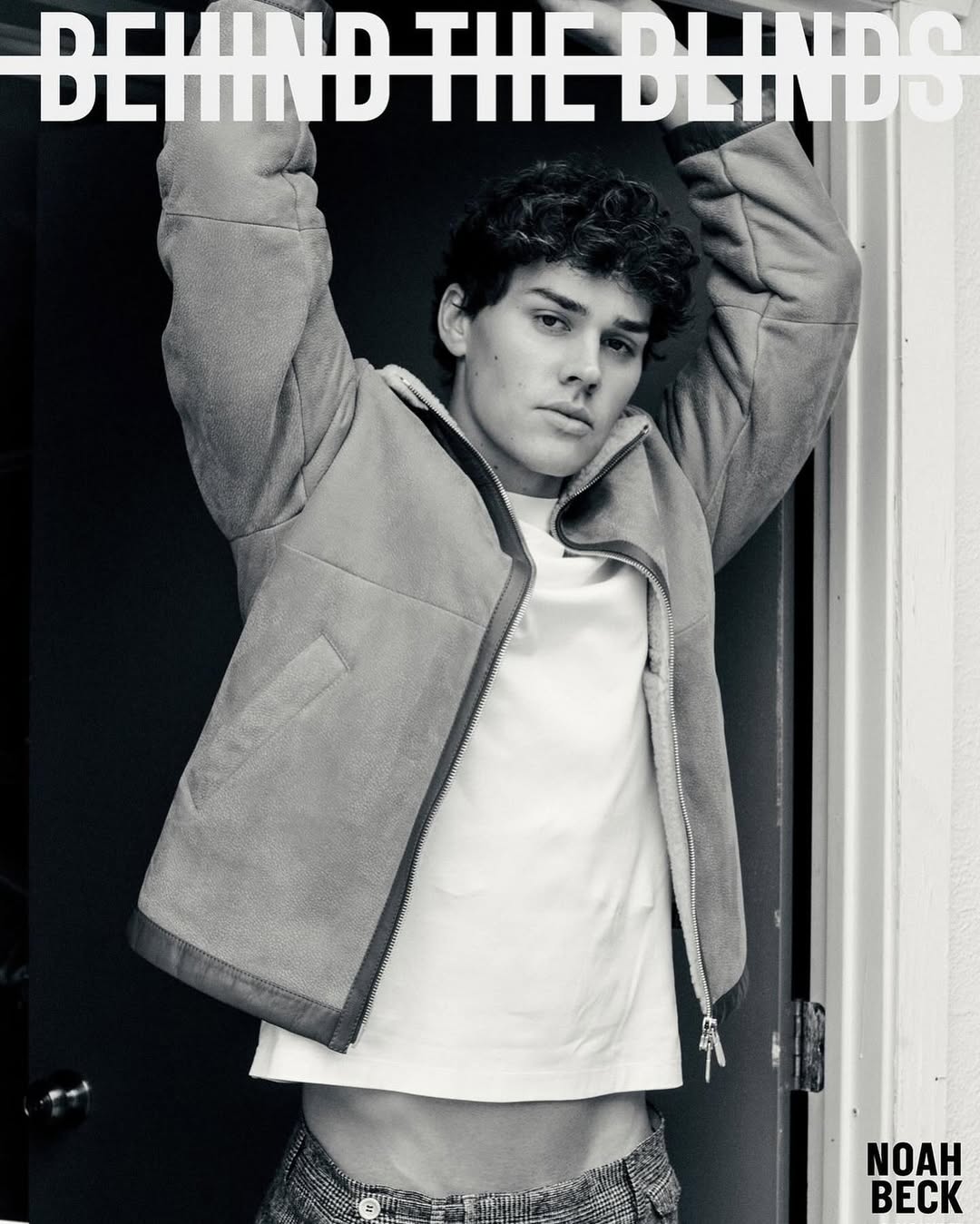 Monochrome studio portrait of a young adult male model wearing a jacket and T-shirt, posing with raised arms near blinds and a doorway.
