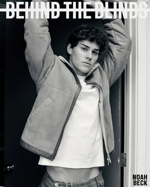 Monochrome studio portrait of a young adult male model wearing a jacket and T-shirt, posing with raised arms near blinds and a doorway.