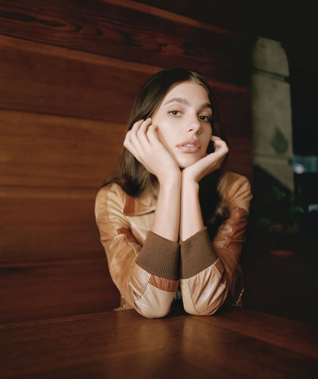 Portrait of a young woman with her hands near her face, wearing a tan jacket, and sitting against a wood background indoors.