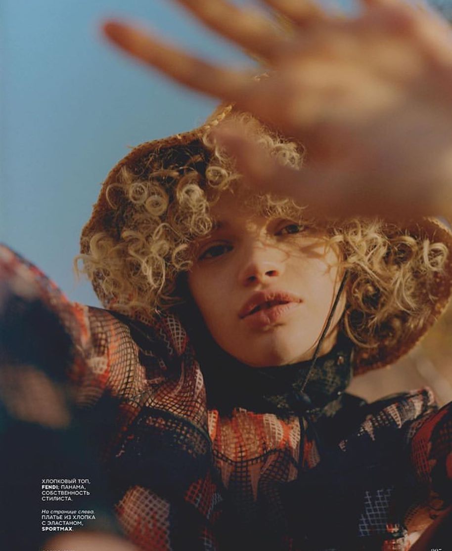Woman poses in a patterned top and hat against a blue sky, partially obscuring her face with her hand in an editorial fashion shot.
