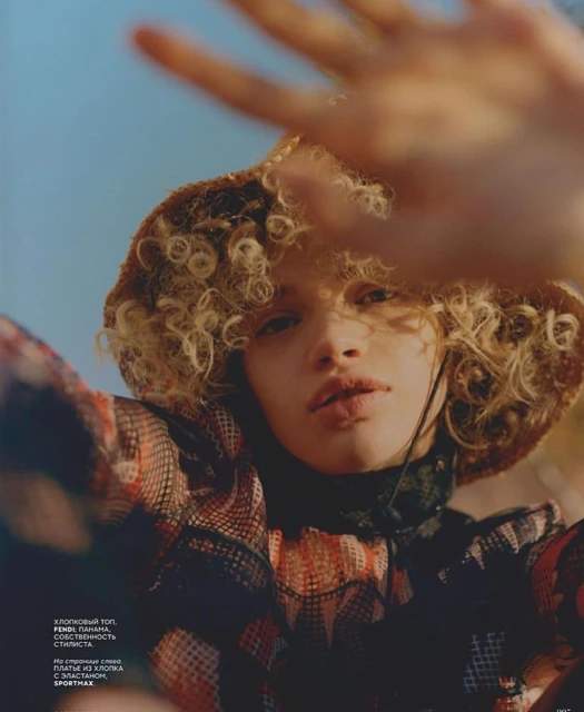 Woman poses in a patterned top and hat against a blue sky, partially obscuring her face with her hand in an editorial fashion shot.