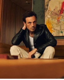 Man with dark hair in a black leather jacket and white tank top sitting on a sofa in a warmly lit room.