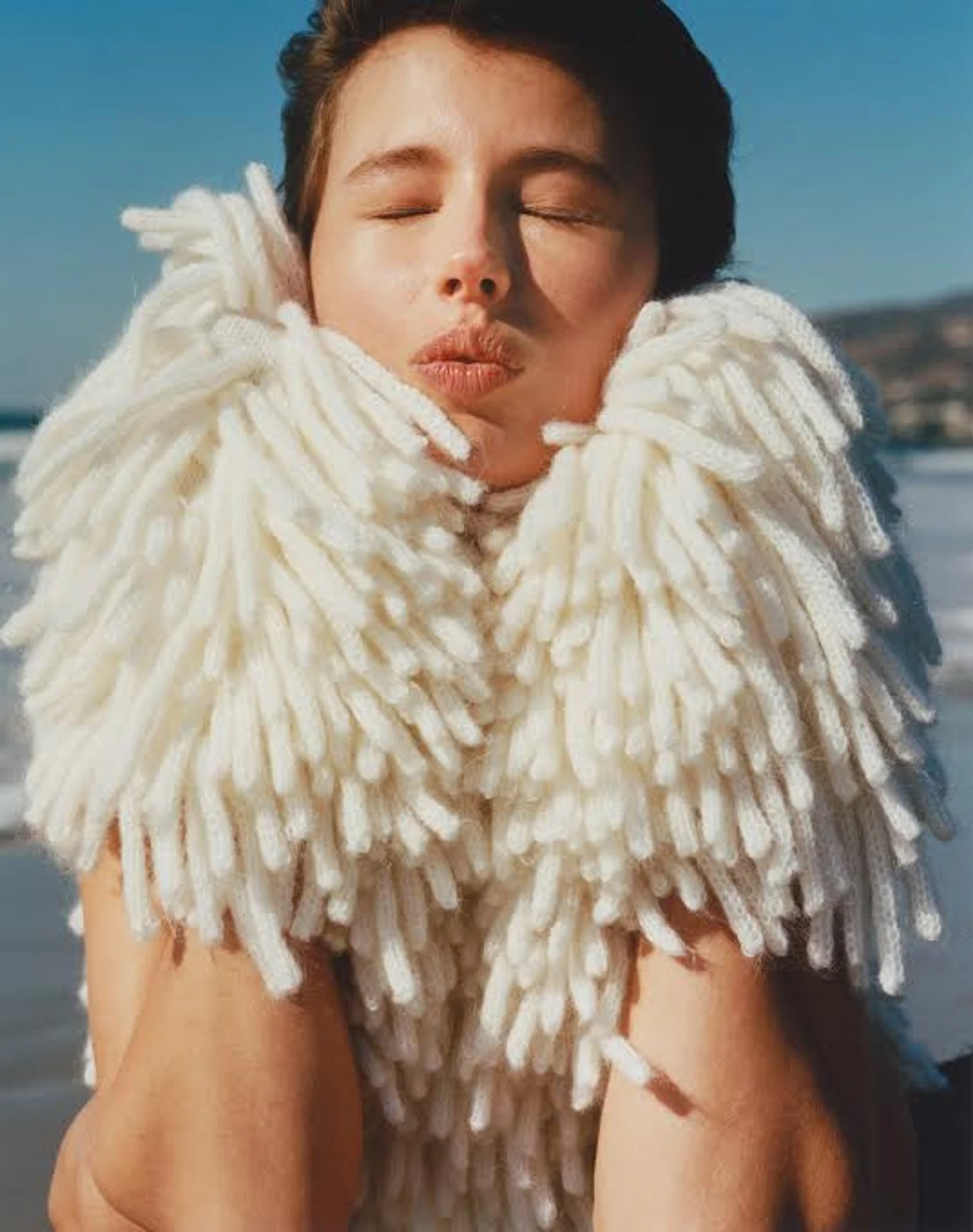 Woman on a beach with eyes closed wearing a large, off-white knitted scarf. The beach is tan with blue water and mountains visible.