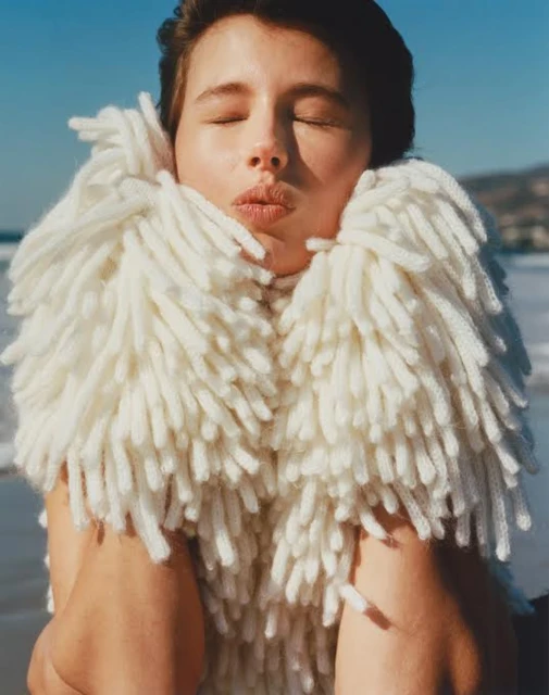 Woman on a beach with eyes closed wearing a large, off-white knitted scarf. The beach is tan with blue water and mountains visible.