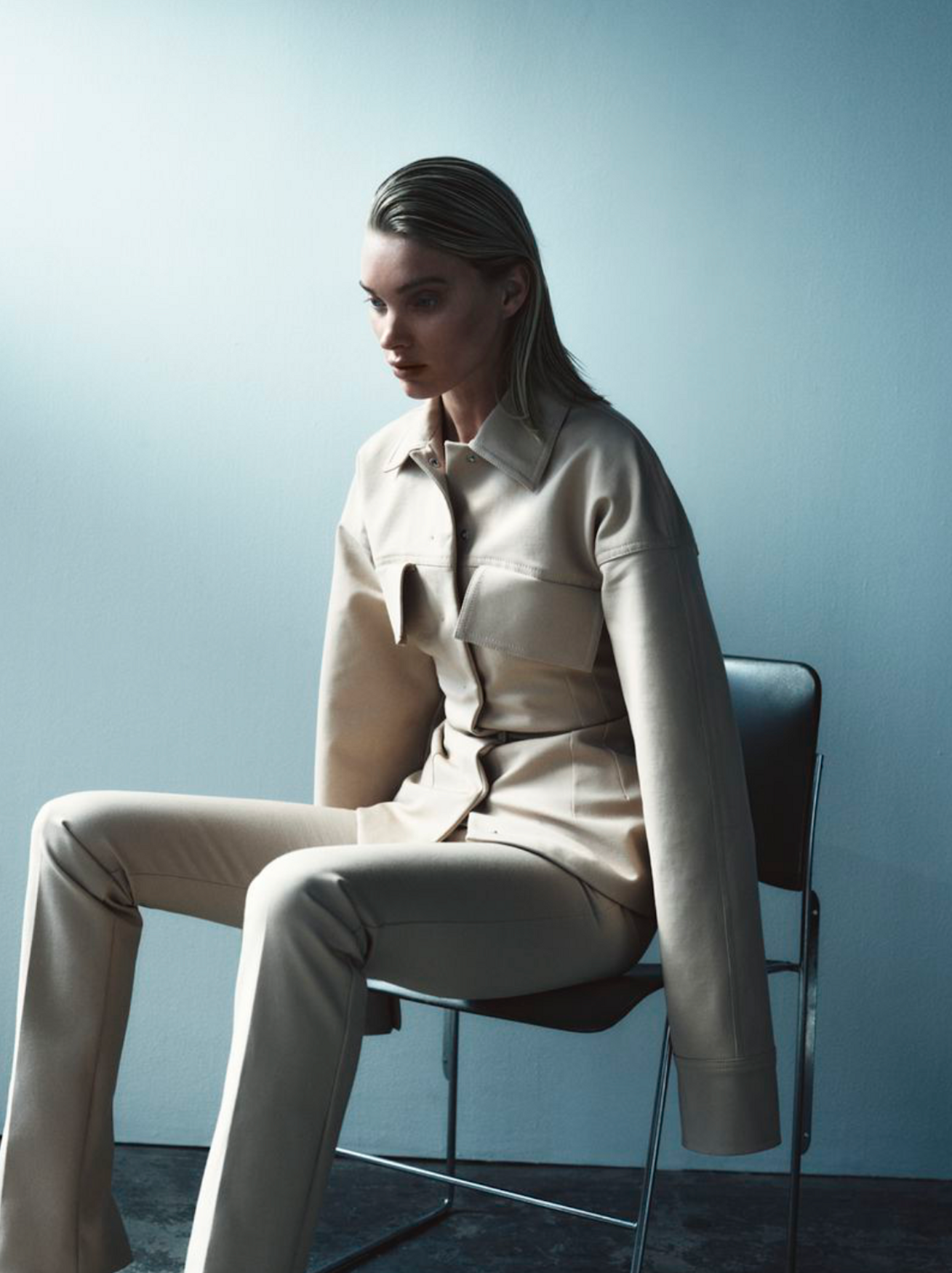 An adult woman in a tan pantsuit sits in a chair against a pale blue background in a studio shot, with wet-look hair.