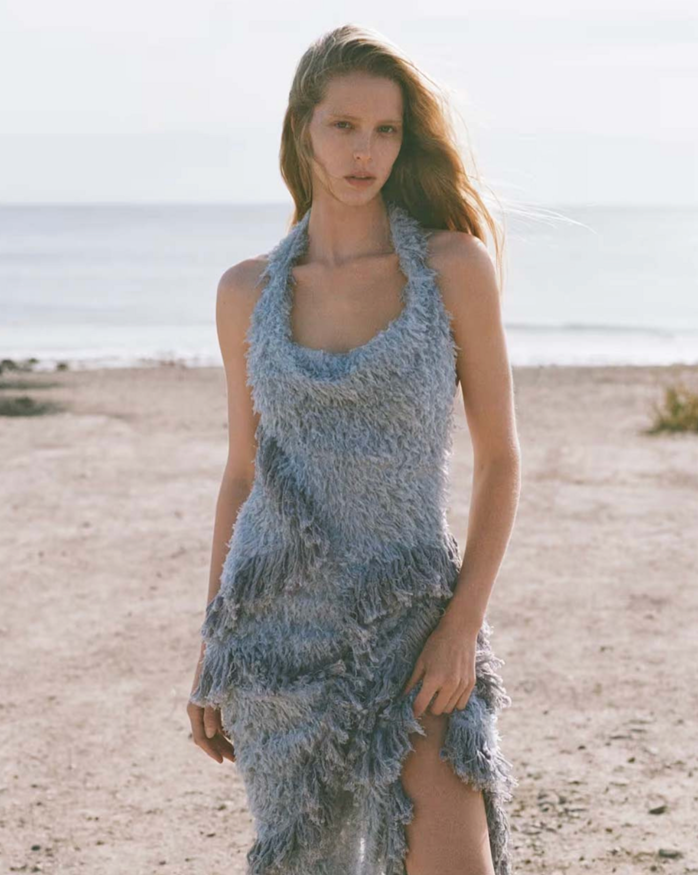 A blonde model poses in a halter dress on a beach with the ocean behind her under an open sky and natural lighting, creating a fashion editorial.